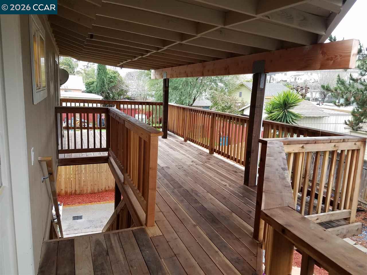 Covered wooden deck with wide plank flooring and railings overlooking neighboring homes.