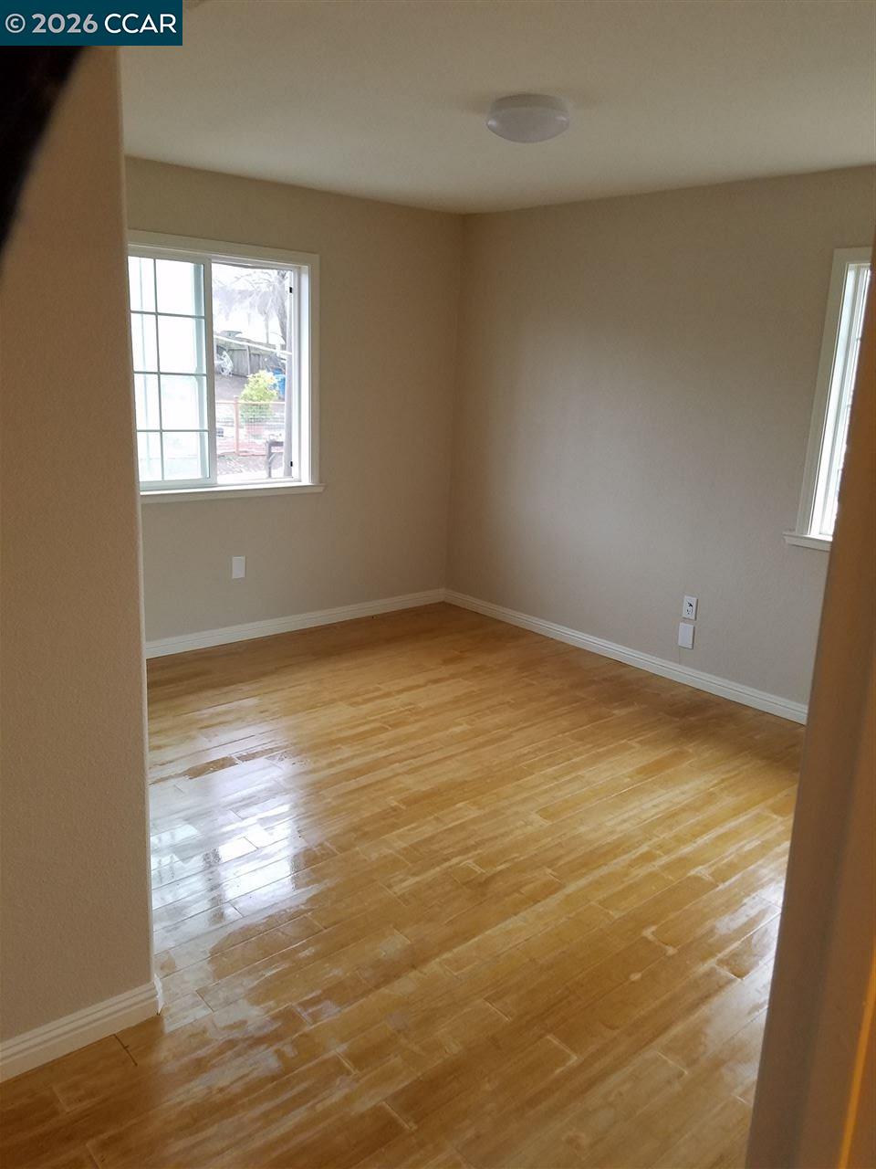 Empty room with glossy hardwood floors, tan walls, and two windows.