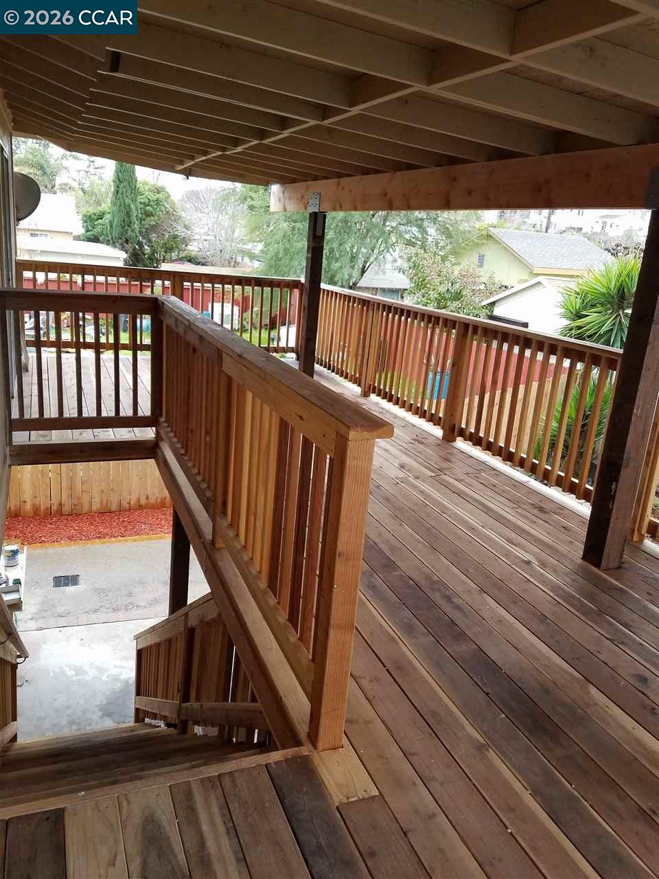 Covered wooden deck with timber railings and stairs overlooking neighboring homes and trees.