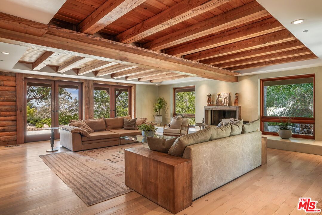 Living room with exposed timber beams, large windows, and sofas arranged around a fireplace.