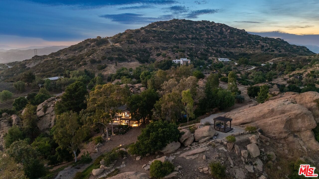 Hillside home glowing at dusk among boulders and trees, with a mountain ridge behind.
