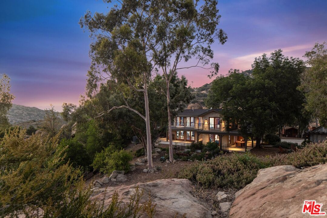 Two-story hillside home with a balcony, mature trees, and rocky landscaping at dusk.