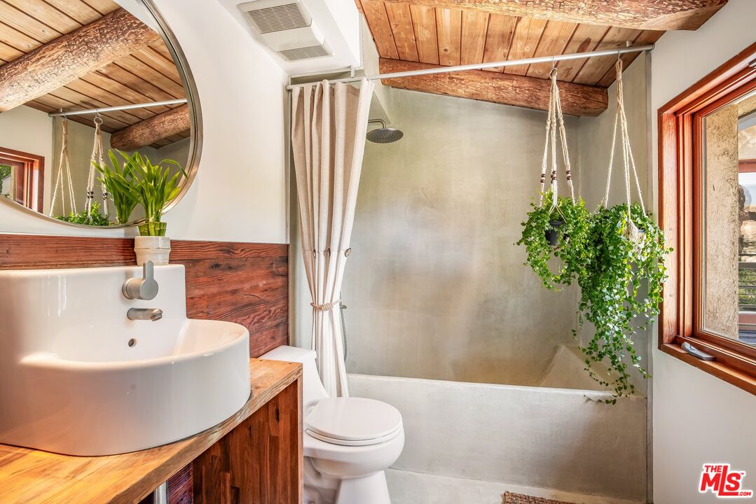 Bathroom with a concrete shower, wood vanity, exposed ceiling beams, and hanging greenery.