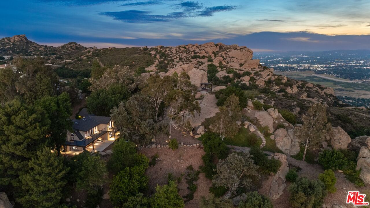 Hillside home among rocky outcrops at dusk, with illuminated windows and distant city lights.