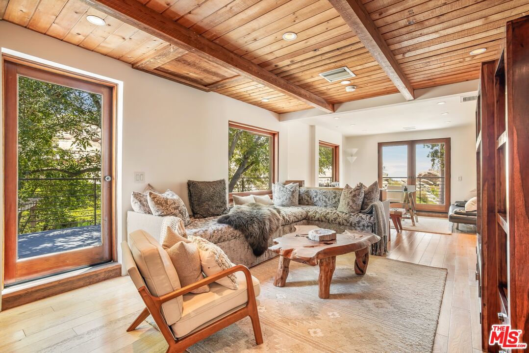 Living room with a vaulted wood ceiling, exposed beams, and large windows opening to a tree-lined deck.