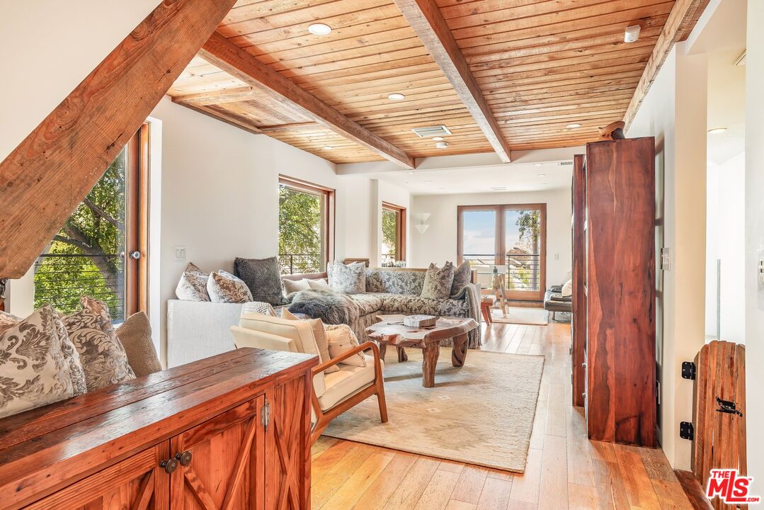 Living room with exposed wood beams, a plank ceiling, and large windows opening onto a balcony.
