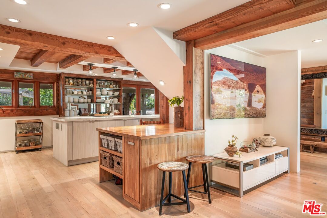 Open-plan kitchen with a wood-topped island, bar stools, exposed beams, and pale wood floors.
