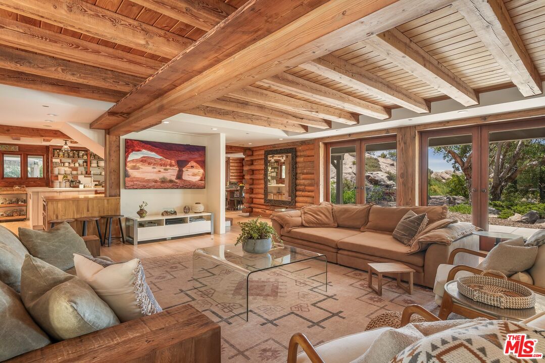 Living room with exposed timber beams, a sectional sofa, and glass doors overlooking a rocky garden.