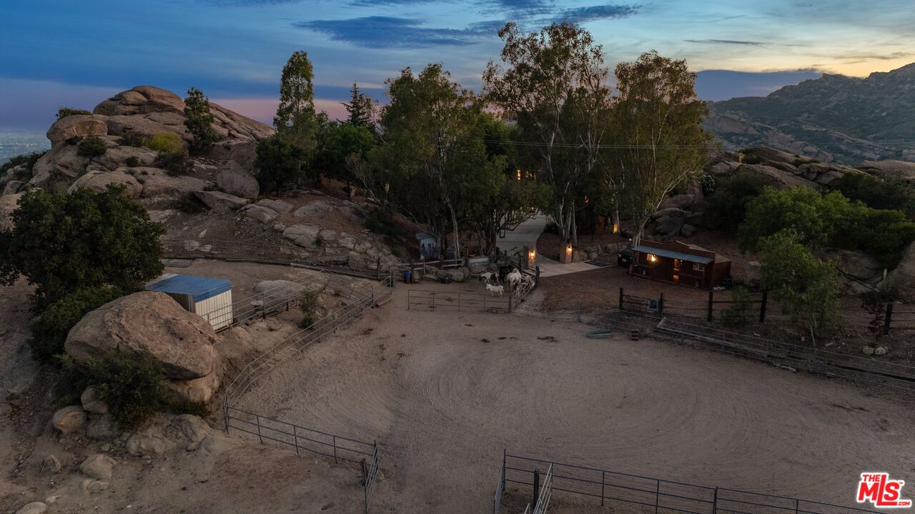 Rocky hillside property with a sandy horse paddock, horses, and outbuildings at dusk.