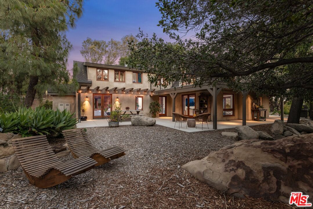 Two-story home with a covered patio, lounge chairs, and mature trees at dusk.