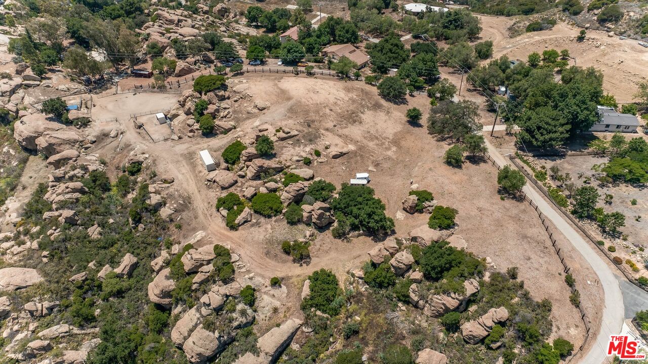 Aerial view of a rocky, dry property with scattered trees, fencing, and neighboring homes.