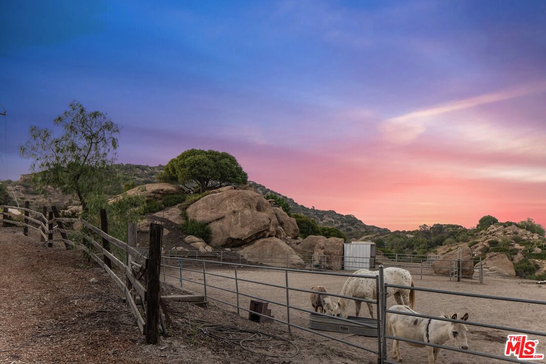 Fenced paddock with two donkeys beside rocky hills beneath a colorful dusk sky.
