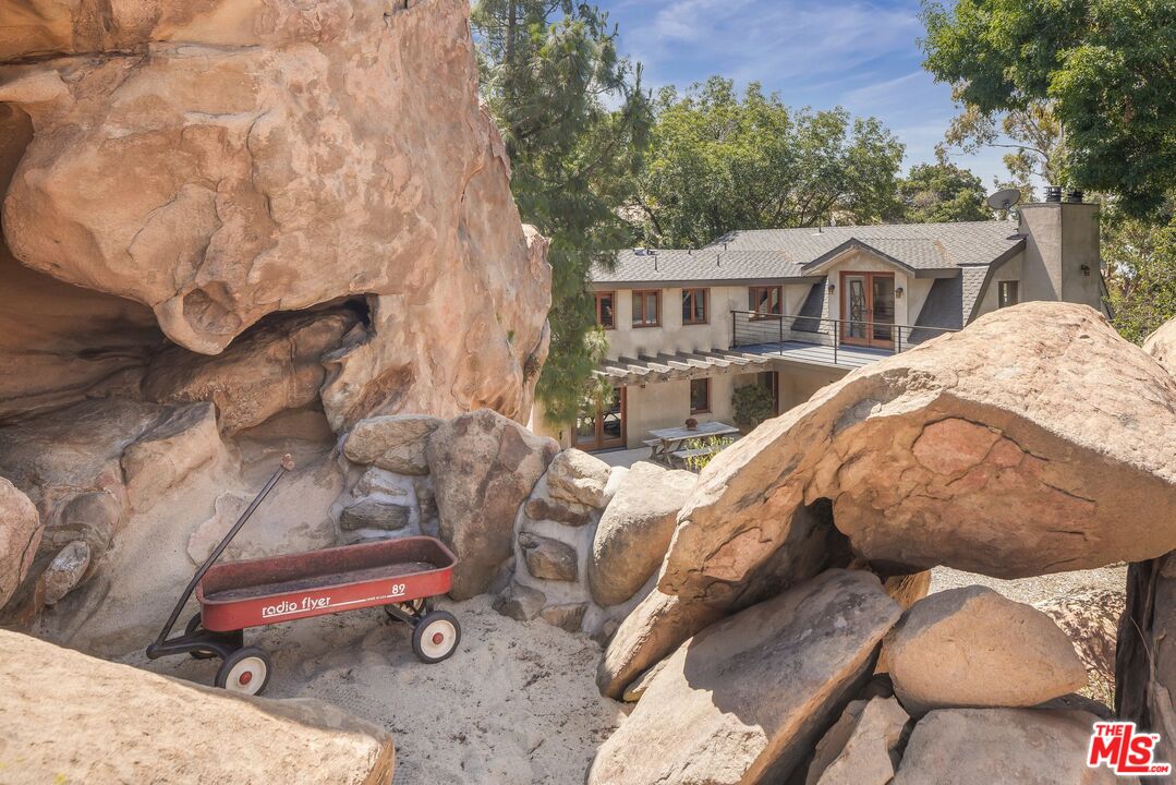 Two-story house framed by sandstone boulders, with a raised deck and red wagon in the sandy foreground.