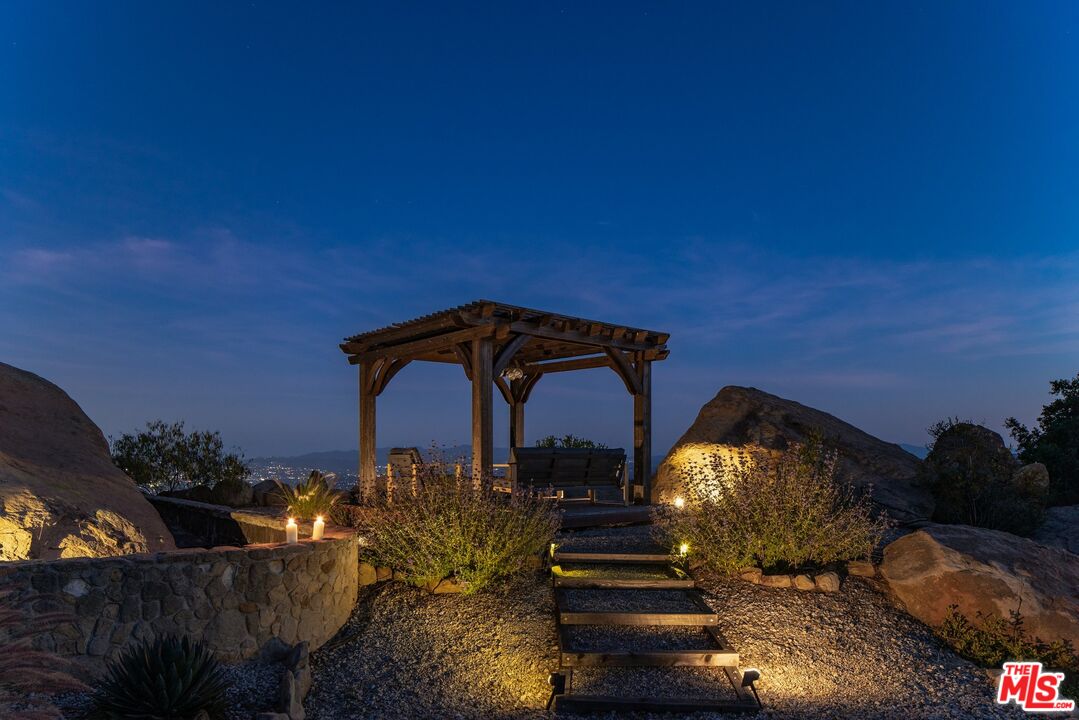 Rocky landscaped yard with a lit wooden pergola and stepping-stone path at dusk.