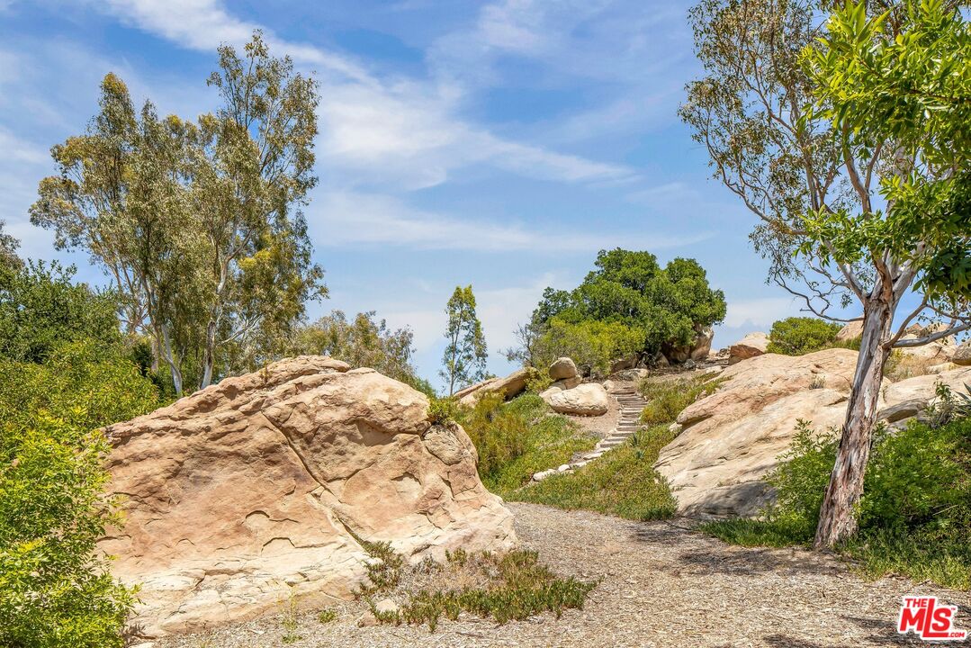 Landscaped grounds with tan boulders, mature trees, and a gravel path leading to stone steps.