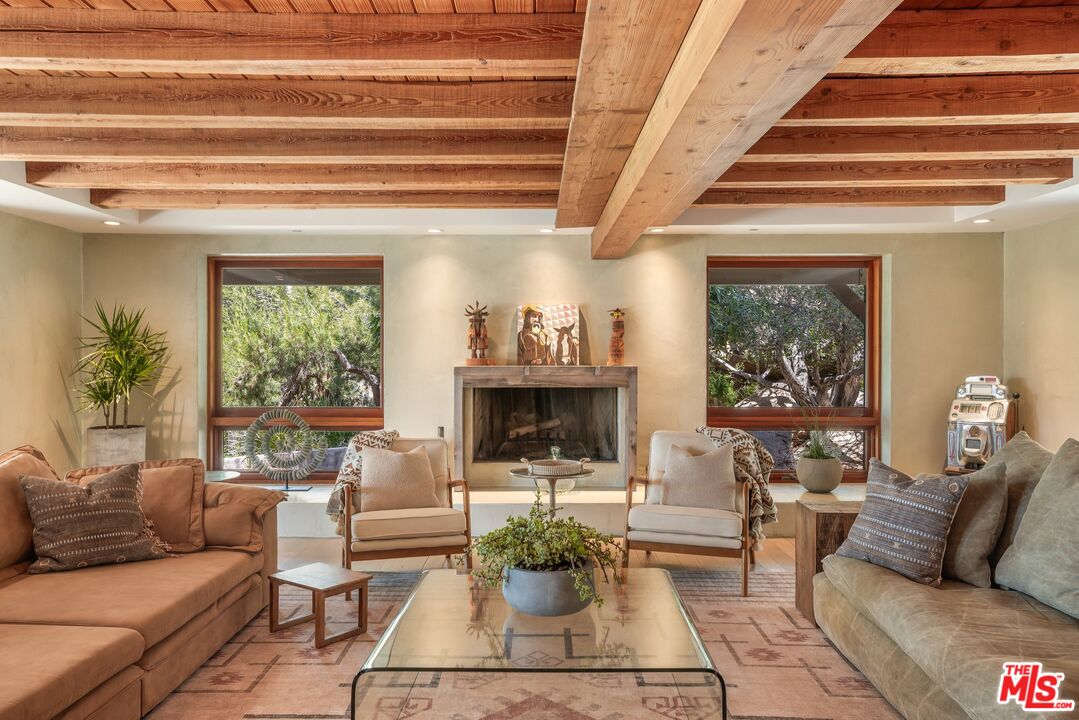 Living room with exposed timber beams, central fireplace, and sofas around a glass coffee table.