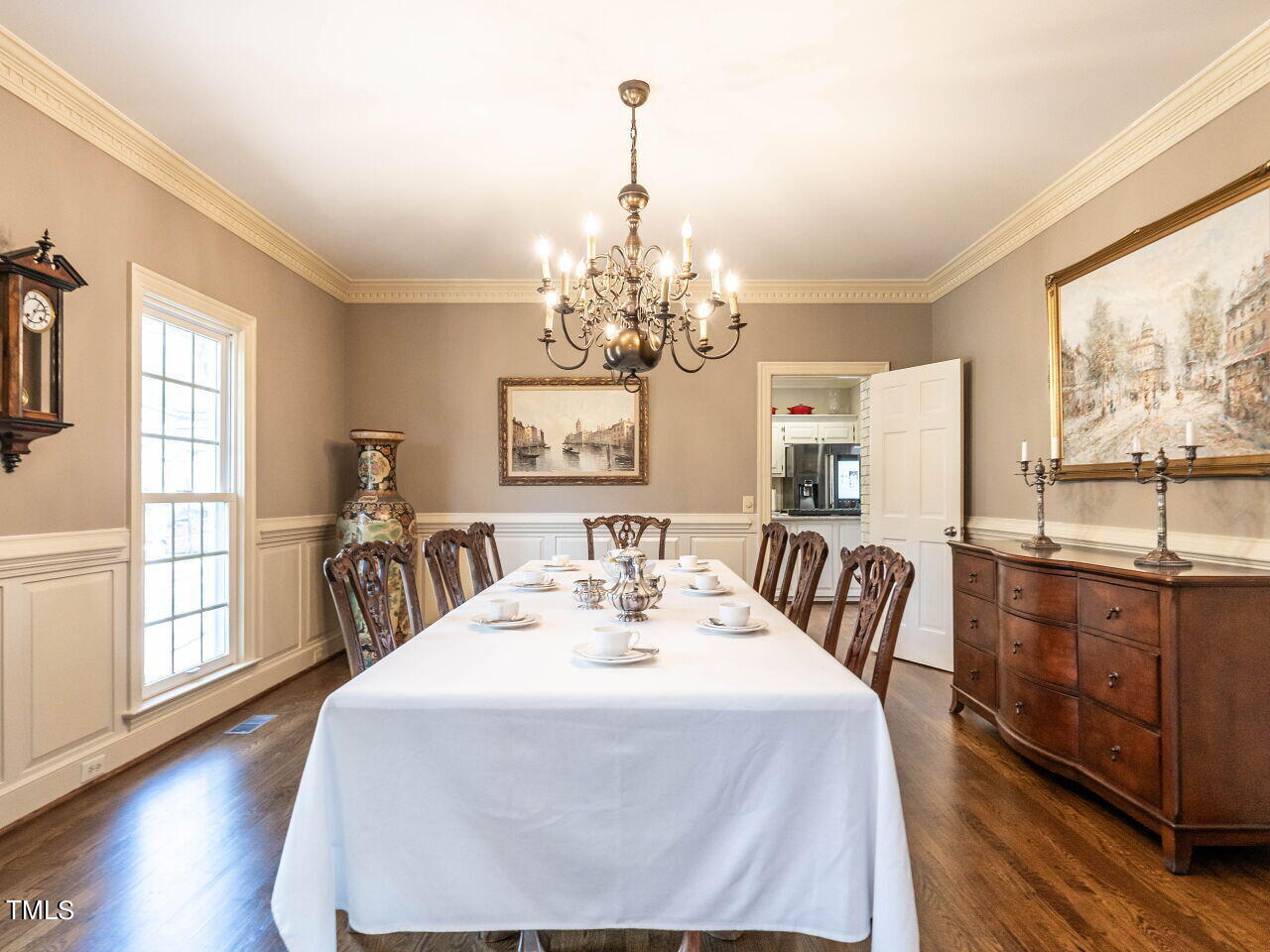 Dining room with a long white-clothed table, carved chairs, chandelier, and hardwood floors.