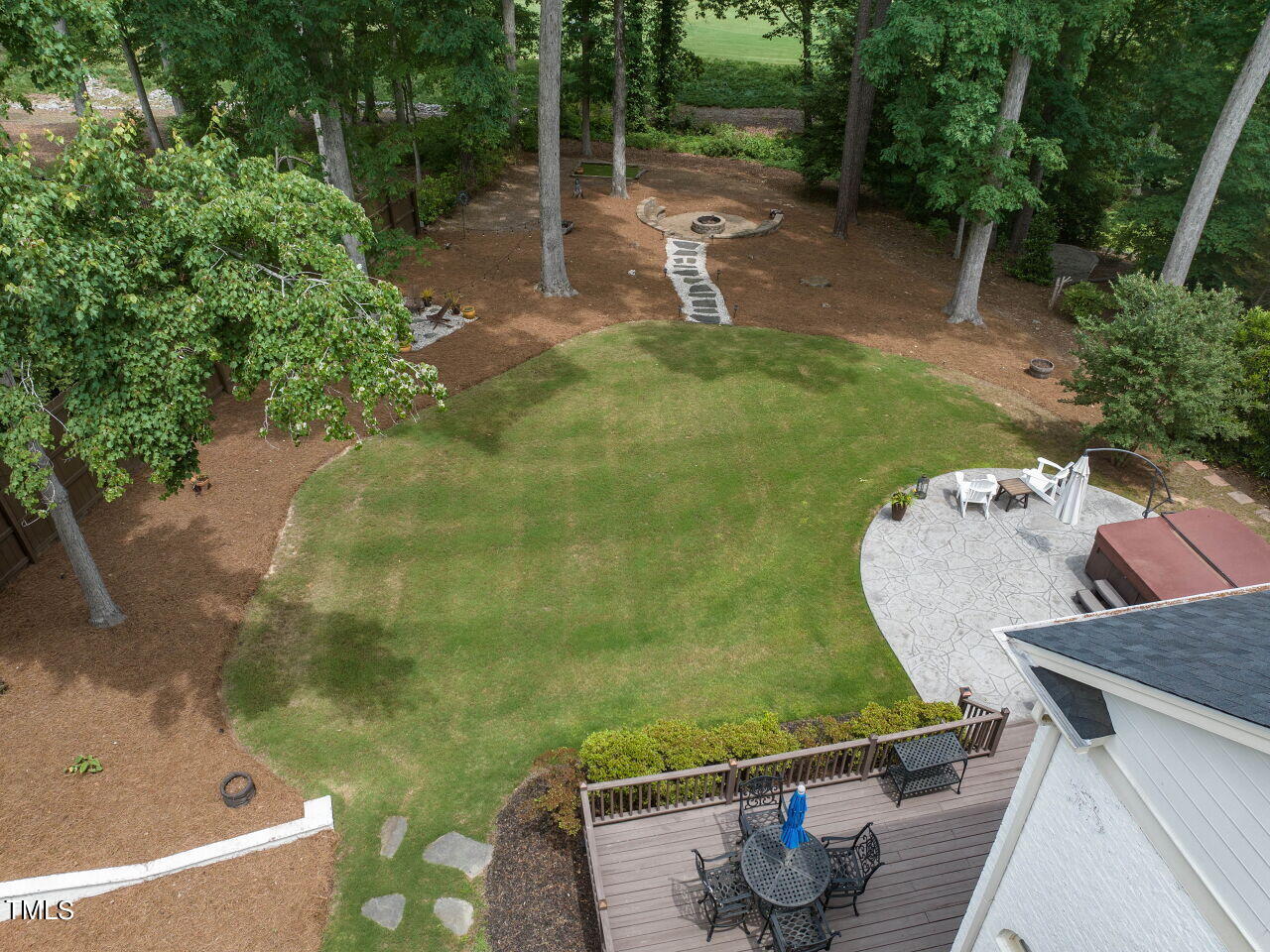 Aerial backyard view with a broad lawn, stone patio, wooden deck, and wooded fire pit area.
