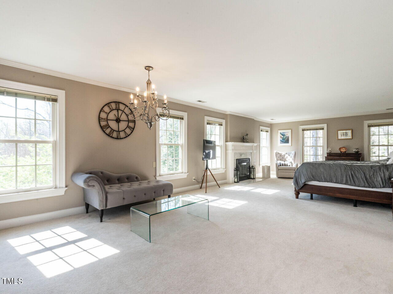 Staged bedroom with a large bed, fireplace, gray chaise lounge, and tall windows.