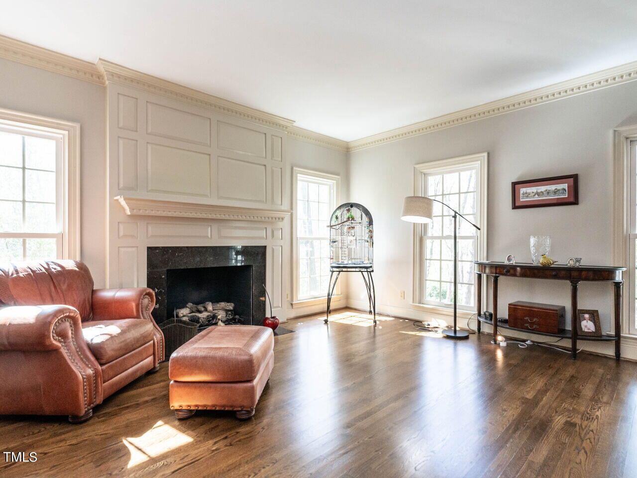 Living room with a paneled fireplace, leather seating, and sunlight across dark hardwood floors.
