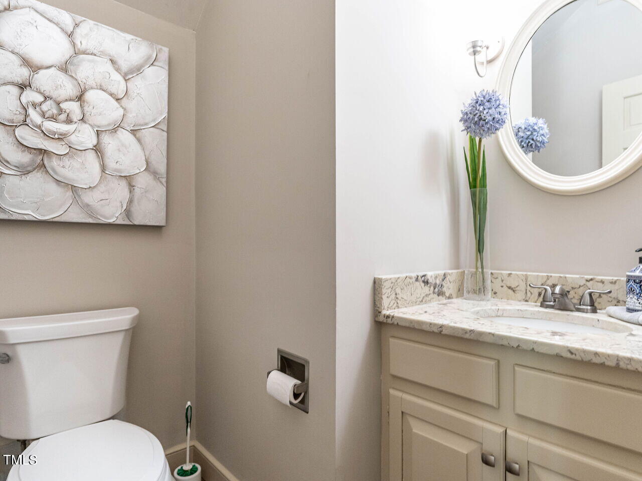 Bathroom with a beige marble-top vanity, oval mirror, blue flowers, and white floral wall art.