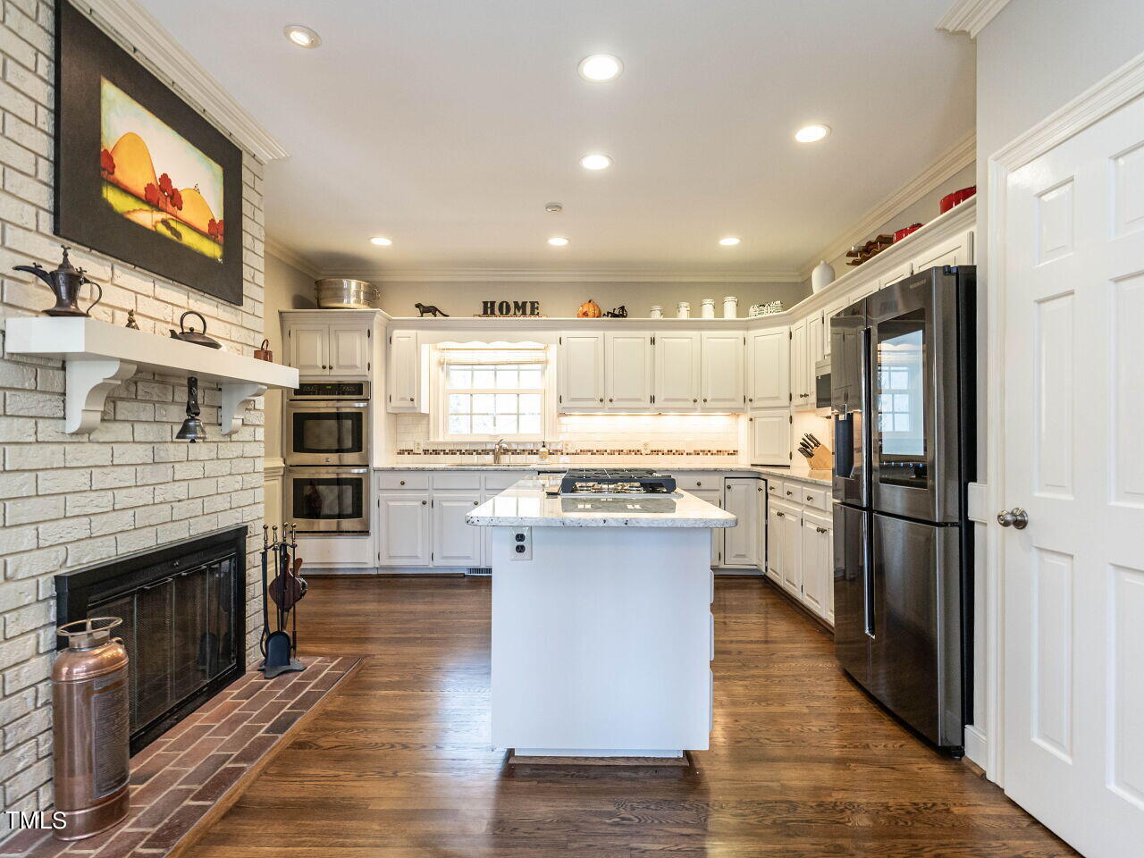 Kitchen with white cabinets, a central cooktop island, stainless steel appliances, and a brick fireplace.