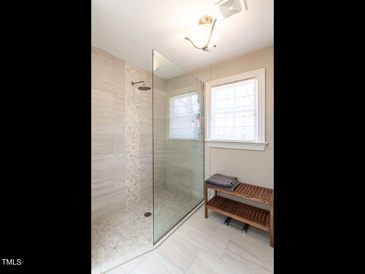 Walk-in shower with glass enclosure, pebble tile accent, and a wooden bench beside a bright window.