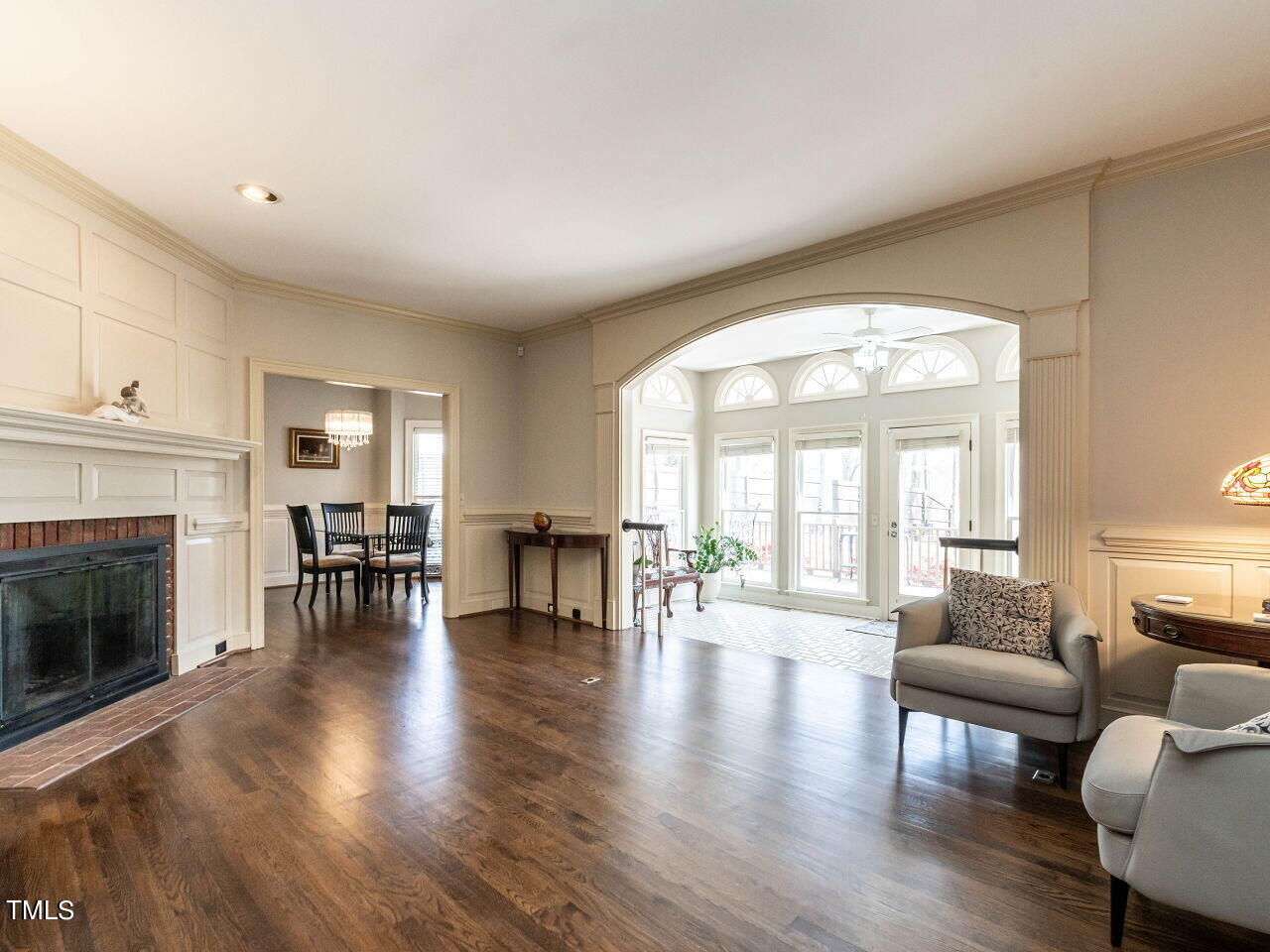 Living room with a paneled fireplace, hardwood floors, and an arched opening to a bright sunroom.