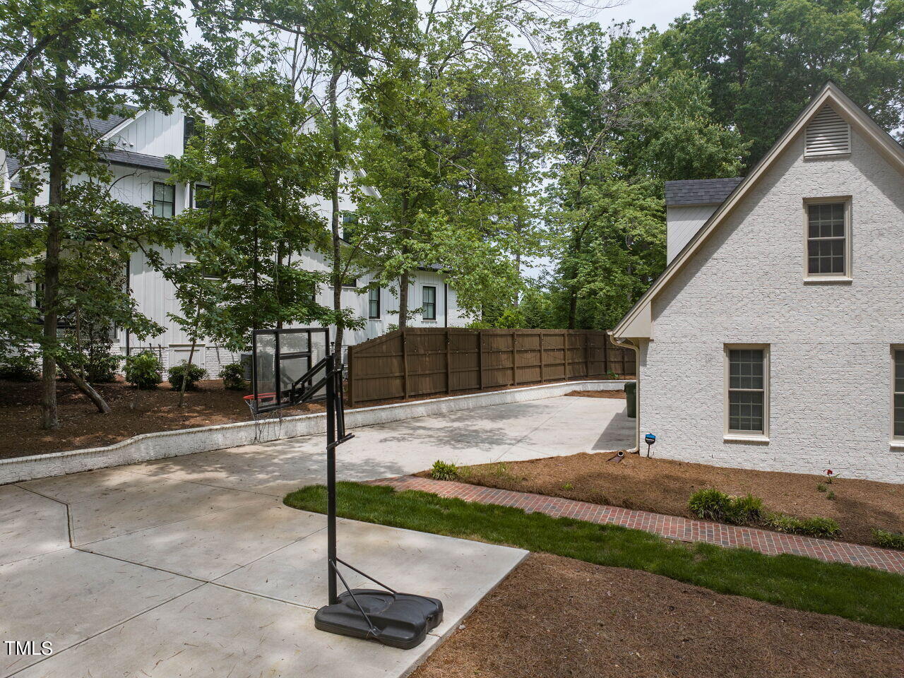 Paved basketball court beside a white house, with a portable hoop and wooden privacy fence.