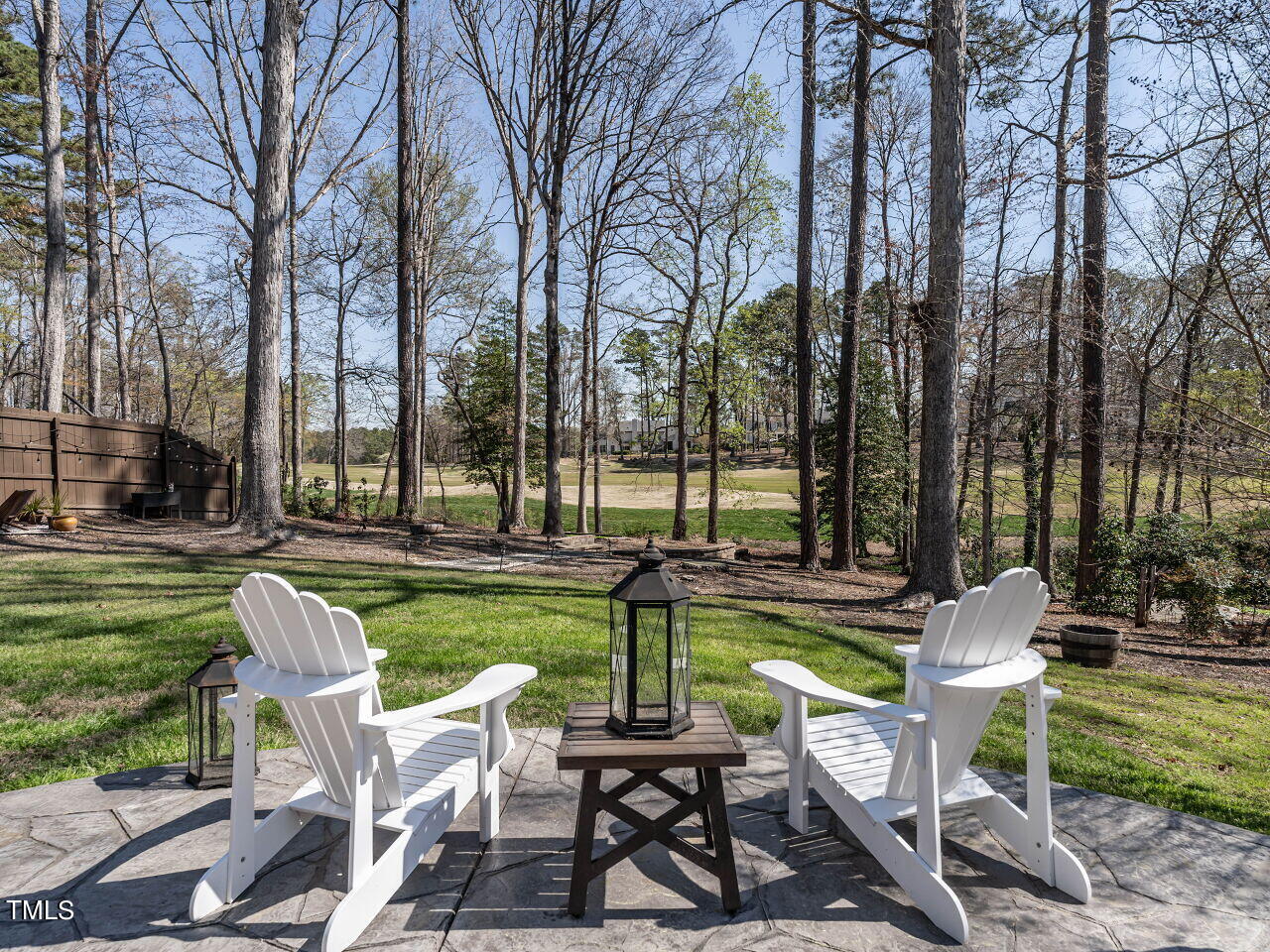 Stone patio with white Adirondack chairs overlooks a wooded backyard and open golf course.