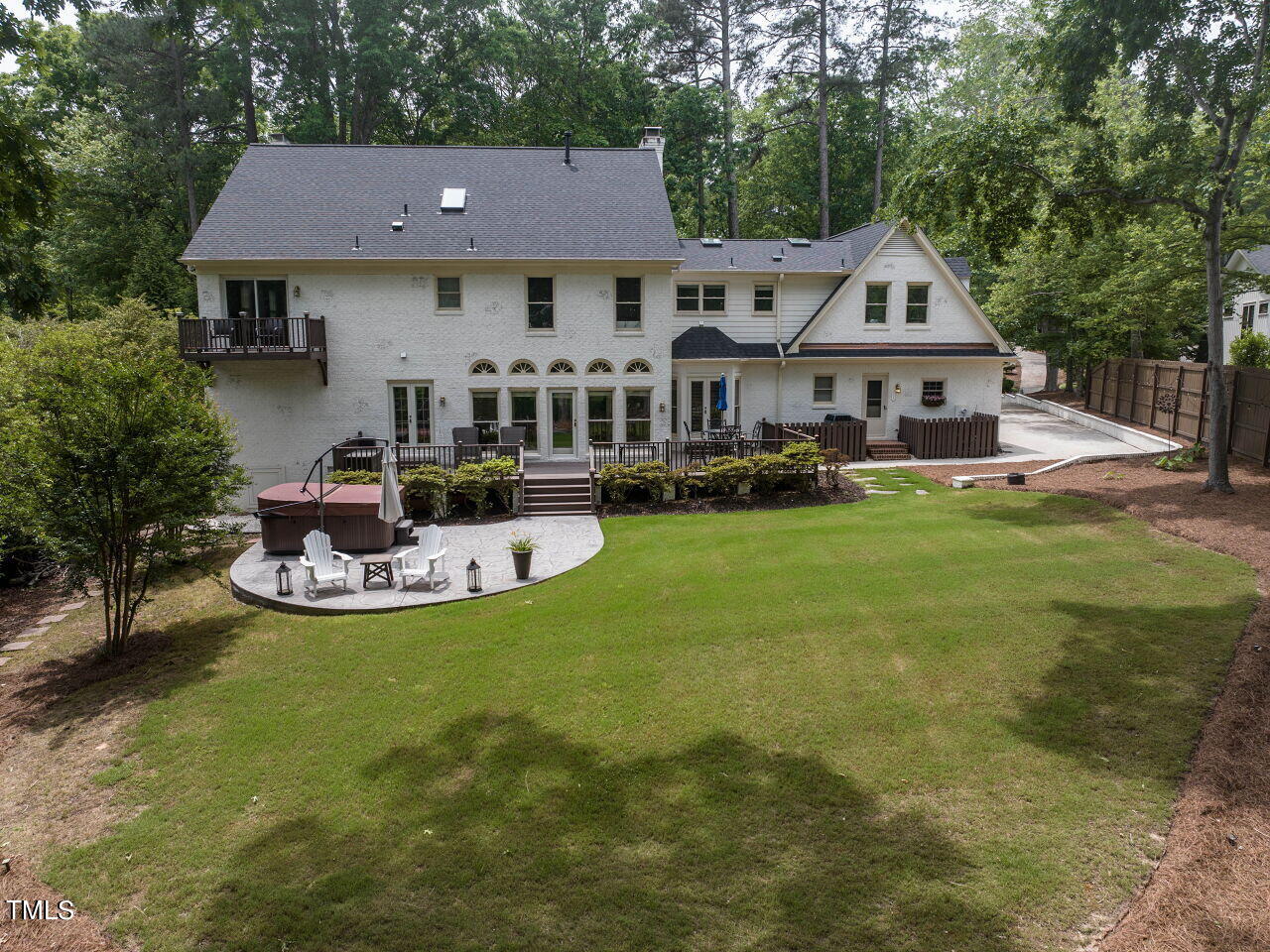 White home's rear exterior with a broad lawn, patio, hot tub, and wooded surroundings.