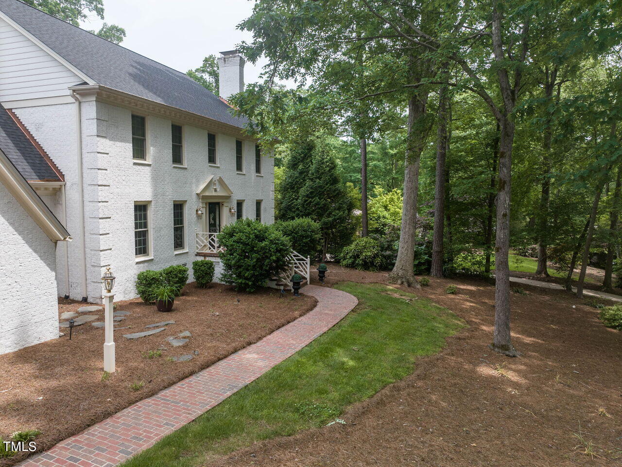 Two-story white brick home with a curved brick walkway beneath mature trees.