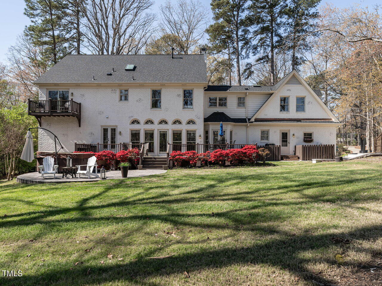 White house with a raised deck, patio seating, red flowering shrubs, and a broad lawn.