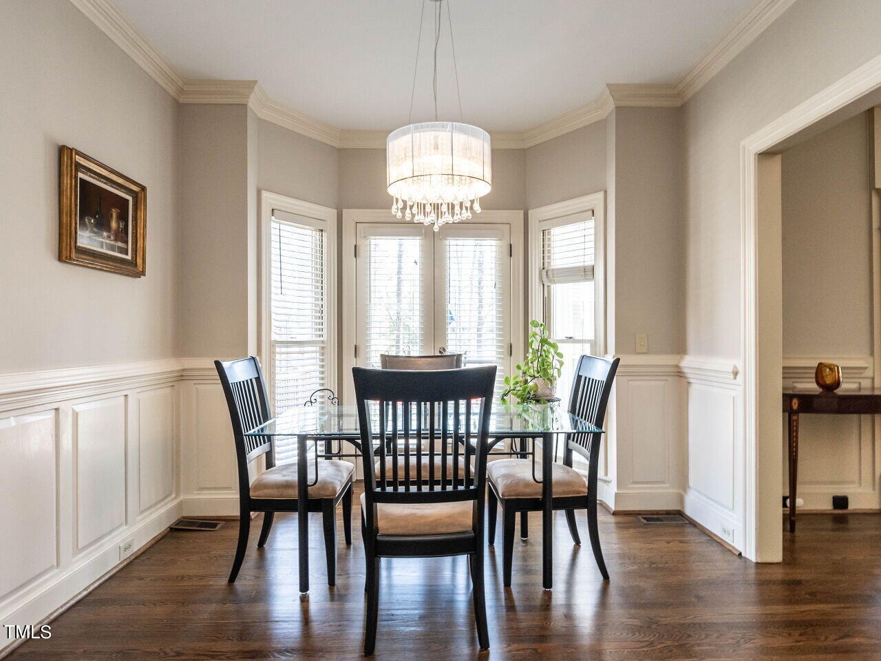 Dining room with a glass-top table, black chairs, and a chandelier beside tall windows.