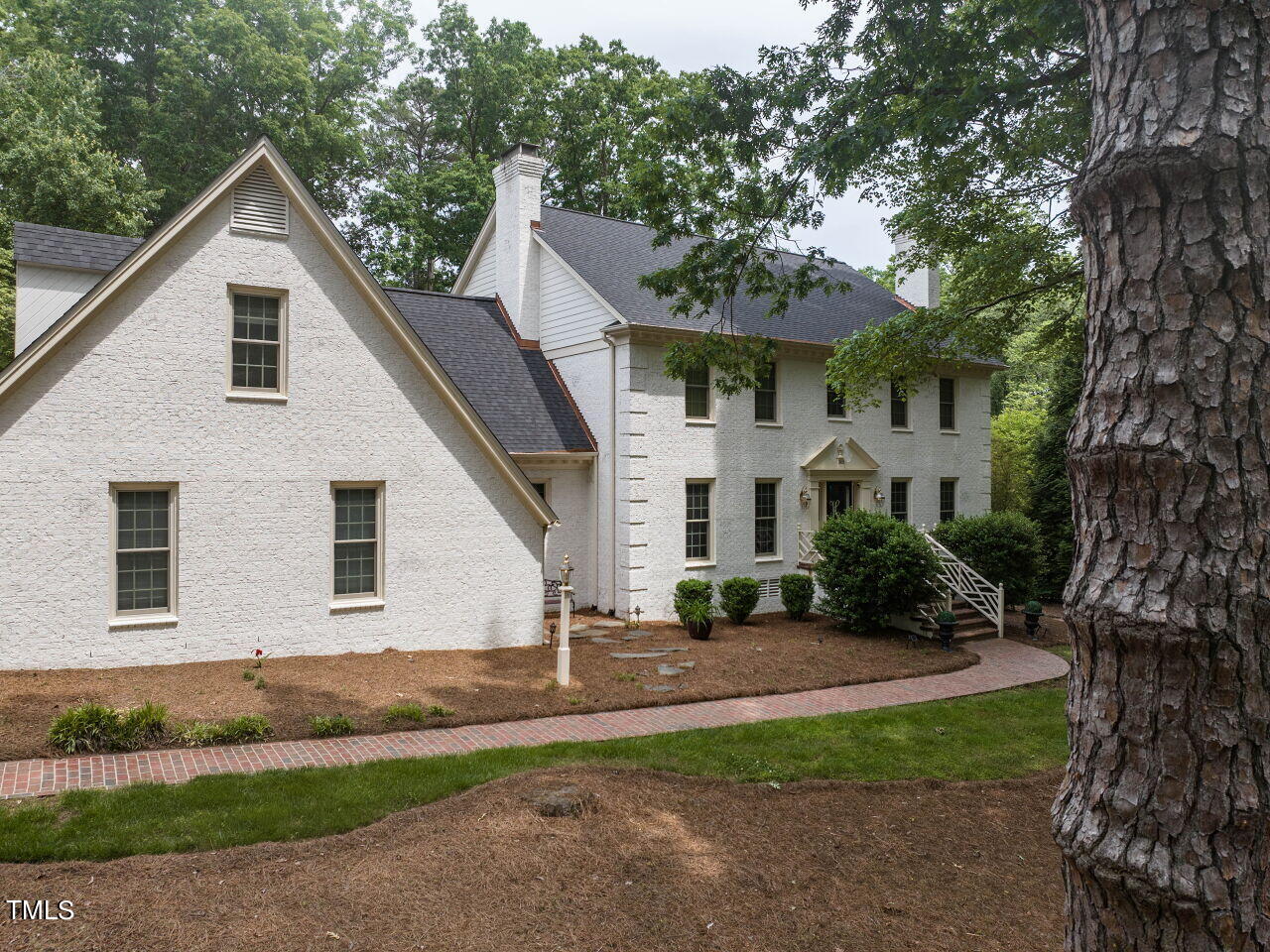 White brick two-story house with steep gables, framed by mature trees and a curved brick walkway.