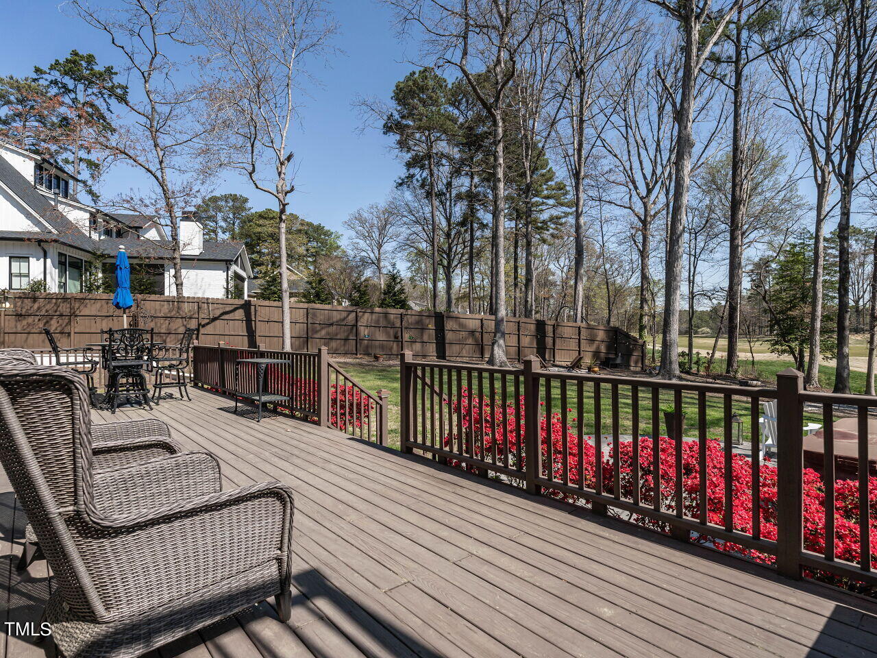 Wooden deck with wicker seating and red flowers overlooking a fenced, tree-filled backyard.