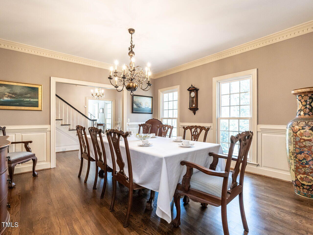 Furnished dining room with a white-clothed table, carved chairs, chandelier, and large windows.