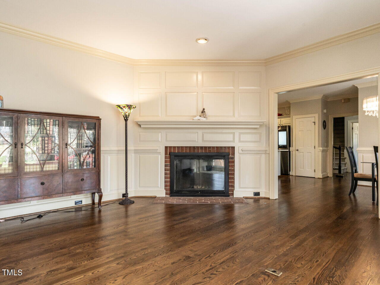 Living room with a brick fireplace, white paneled mantel, hardwood floors, and glass-front wooden cabinet.