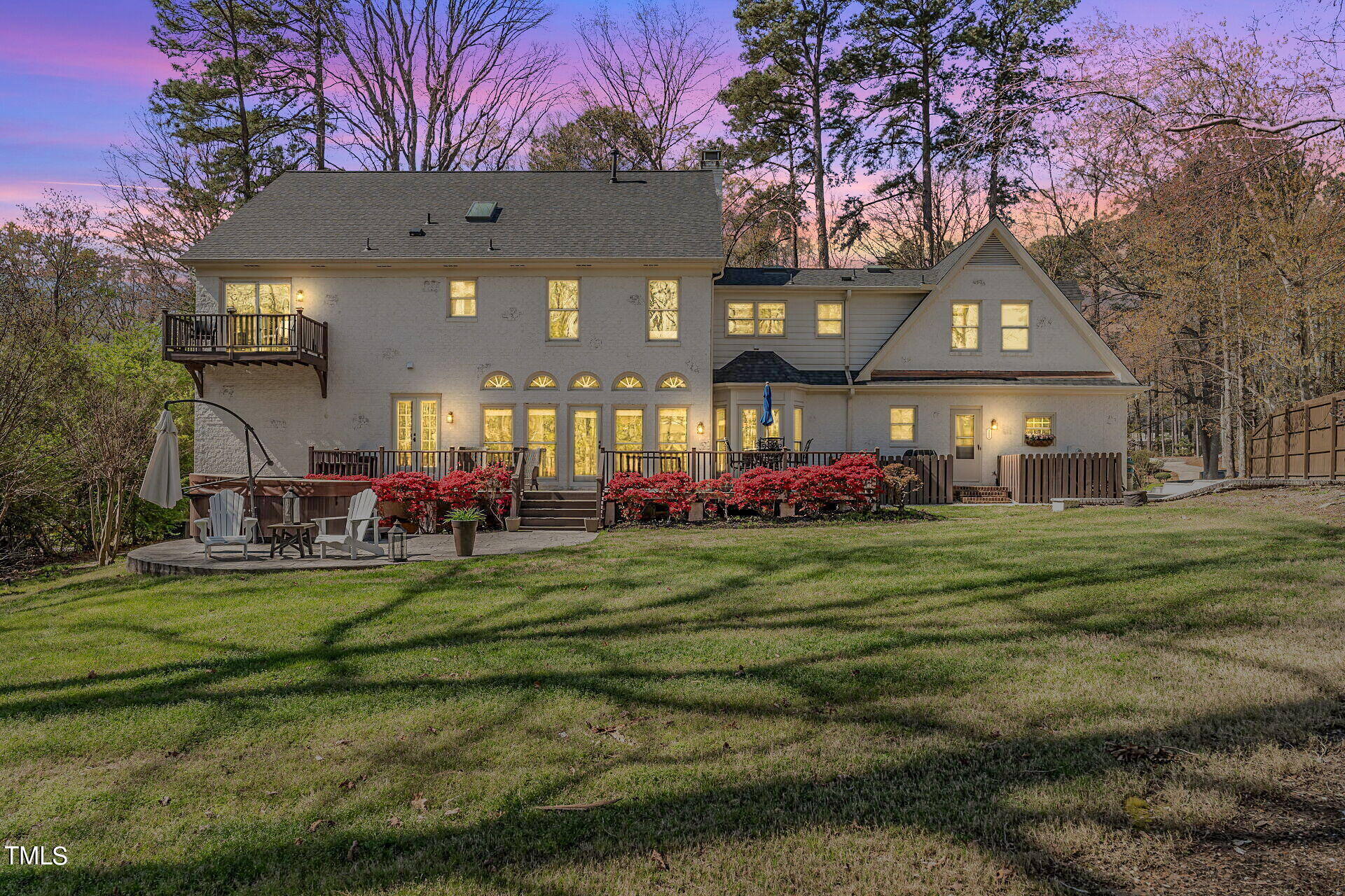 Rear of a large house at dusk, with illuminated windows, a deck, patio seating, and red flowering shrubs.