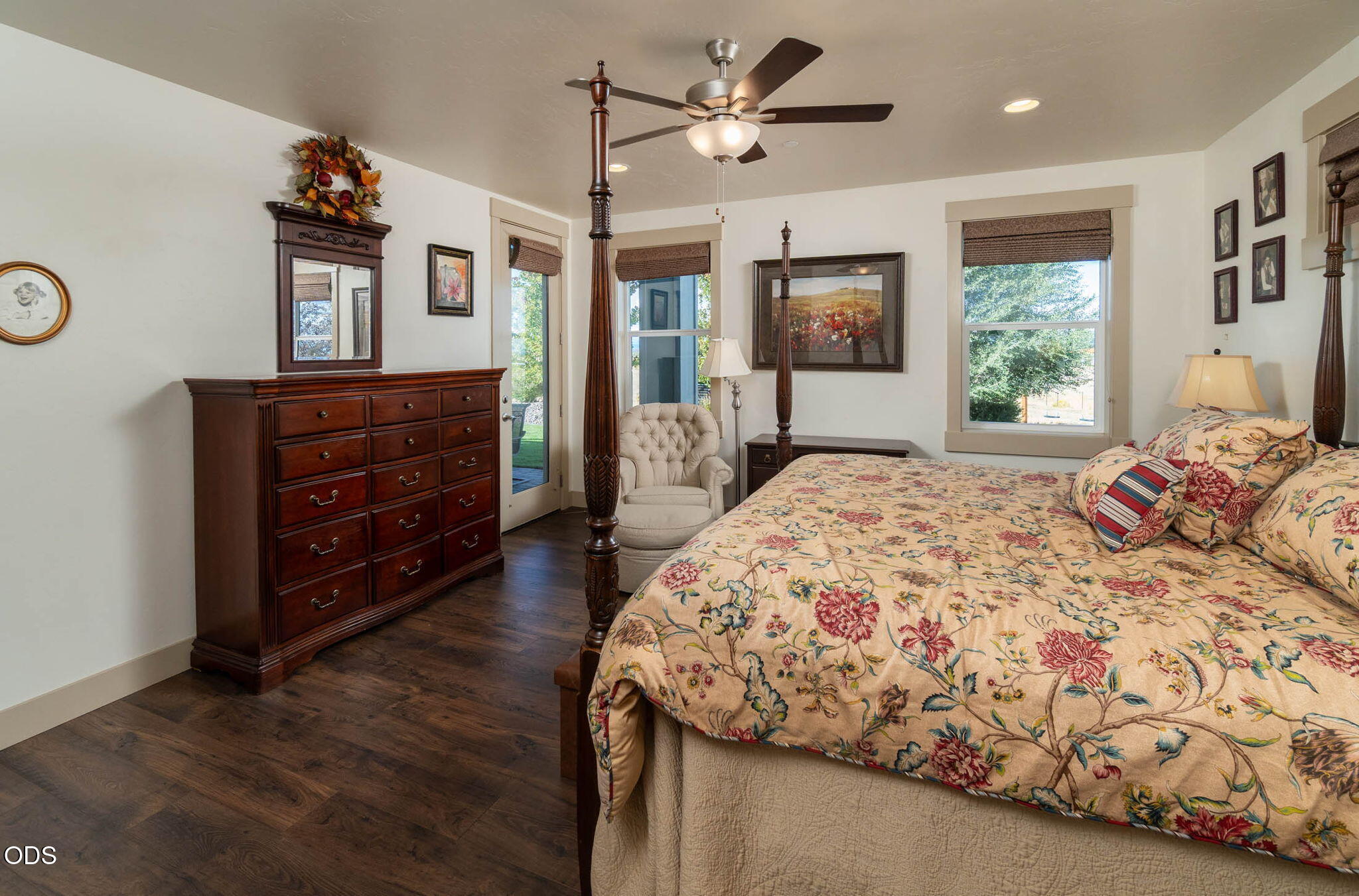 Furnished bedroom with a floral bedspread, dark wood dresser, and windows overlooking green trees.