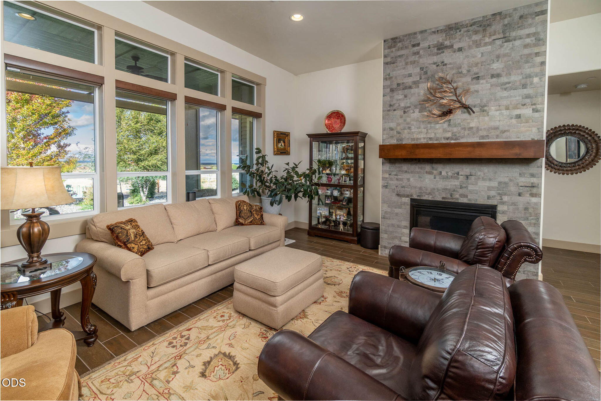 Living room with a stone fireplace, beige sofa, leather chairs, and tall windows.
