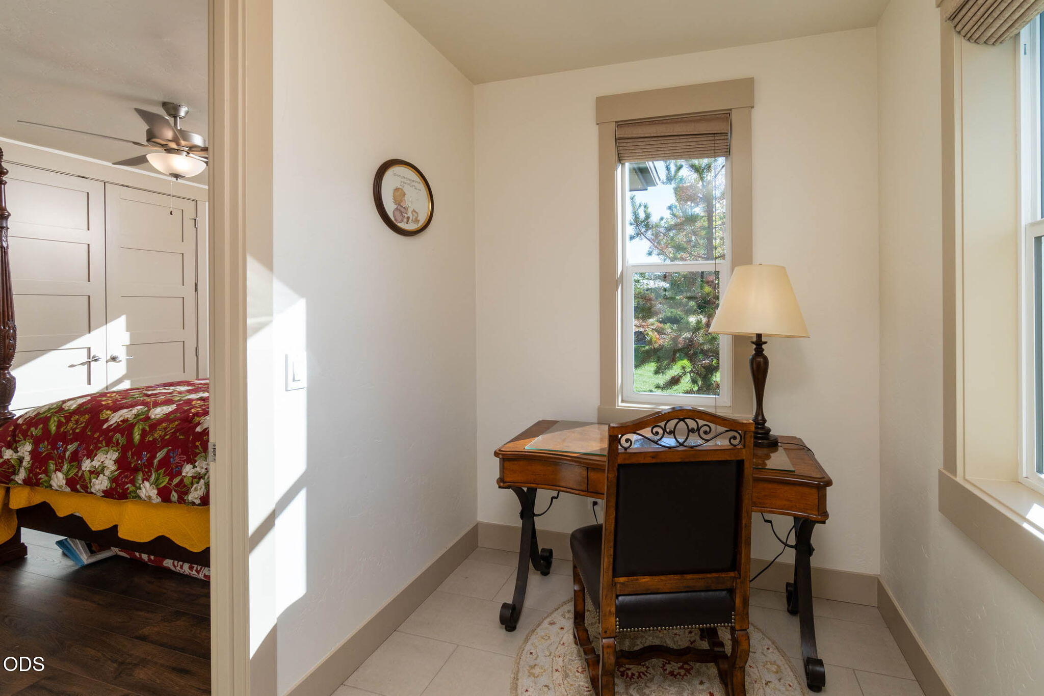 Furnished home office with a wooden desk beside a window, opening onto a bedroom with a floral bedspread.