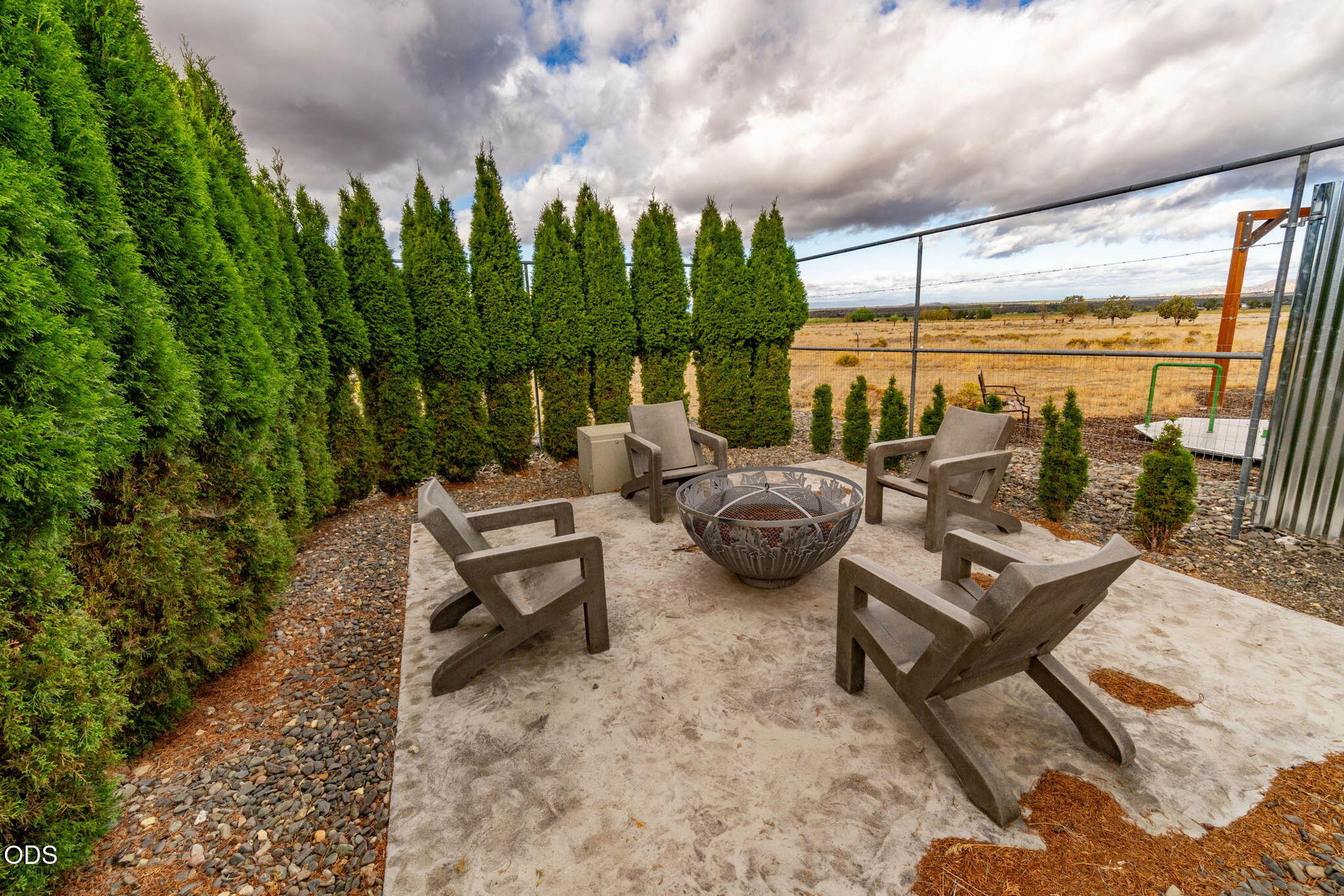 Patio with four chairs around a metal fire pit, bordered by evergreen hedges and open grassland.