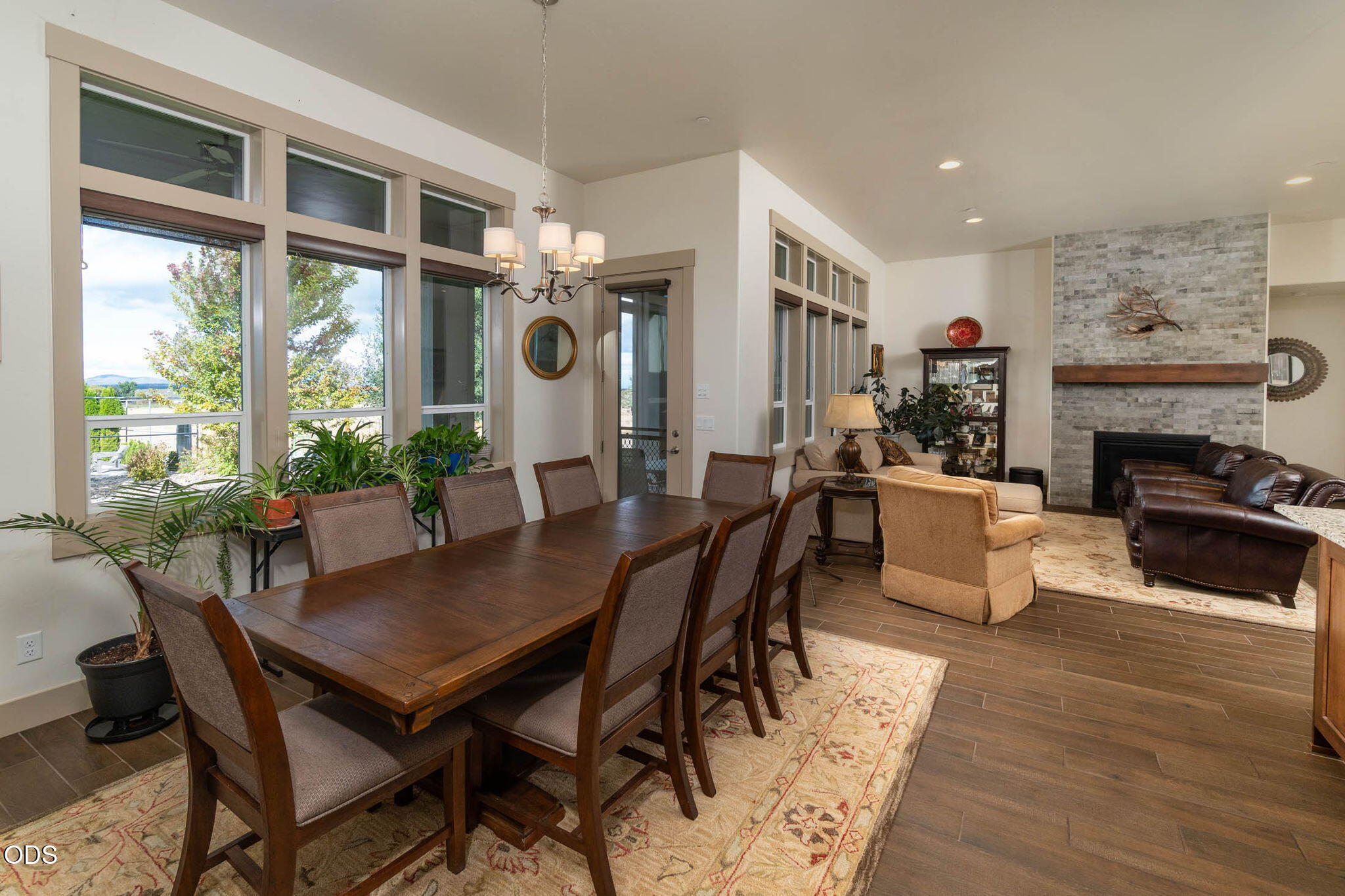 Furnished dining and living room with tall windows, a wood dining table, and a stone fireplace.