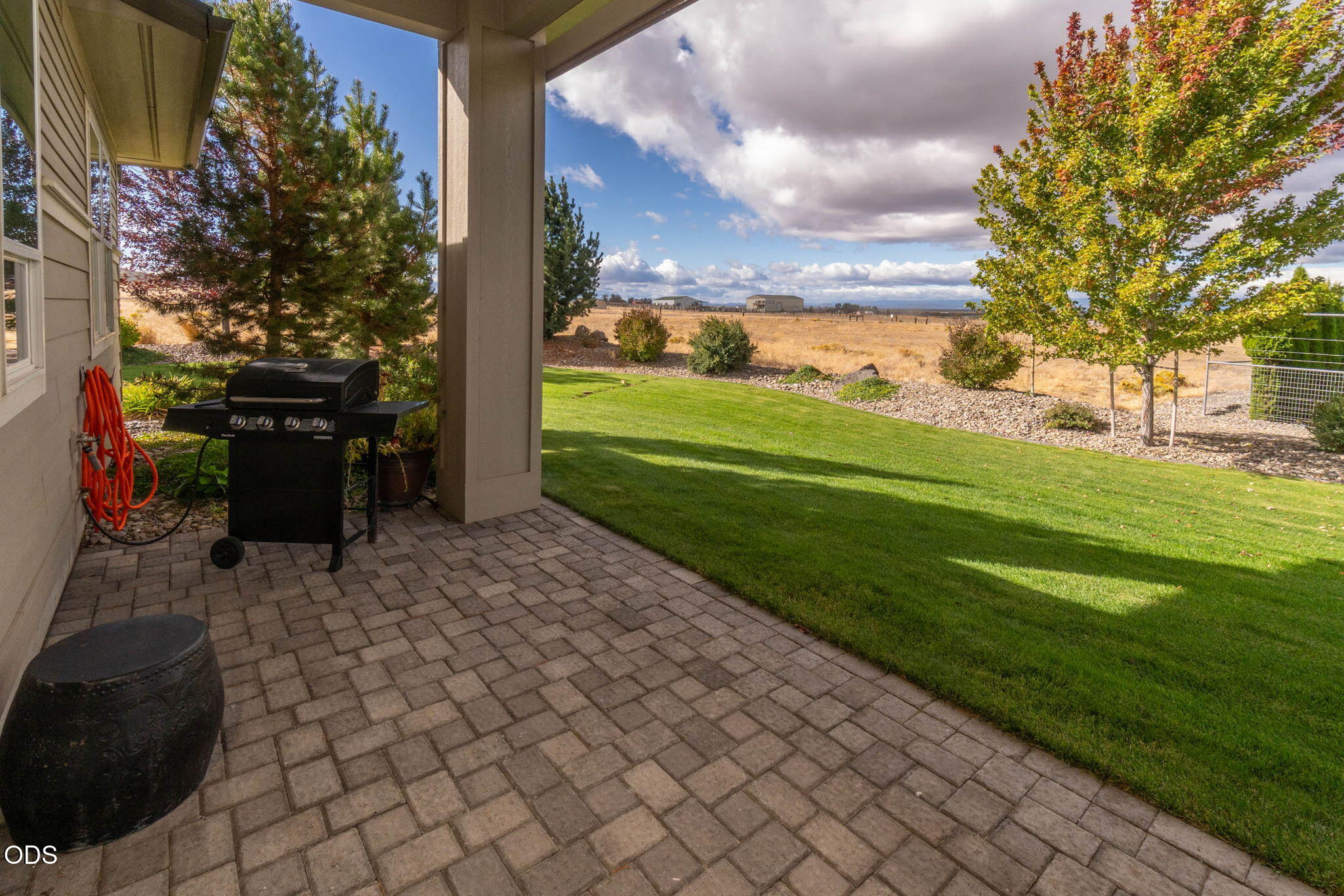 Covered paver patio with a barbecue grill overlooks a lawn and open grassland.
