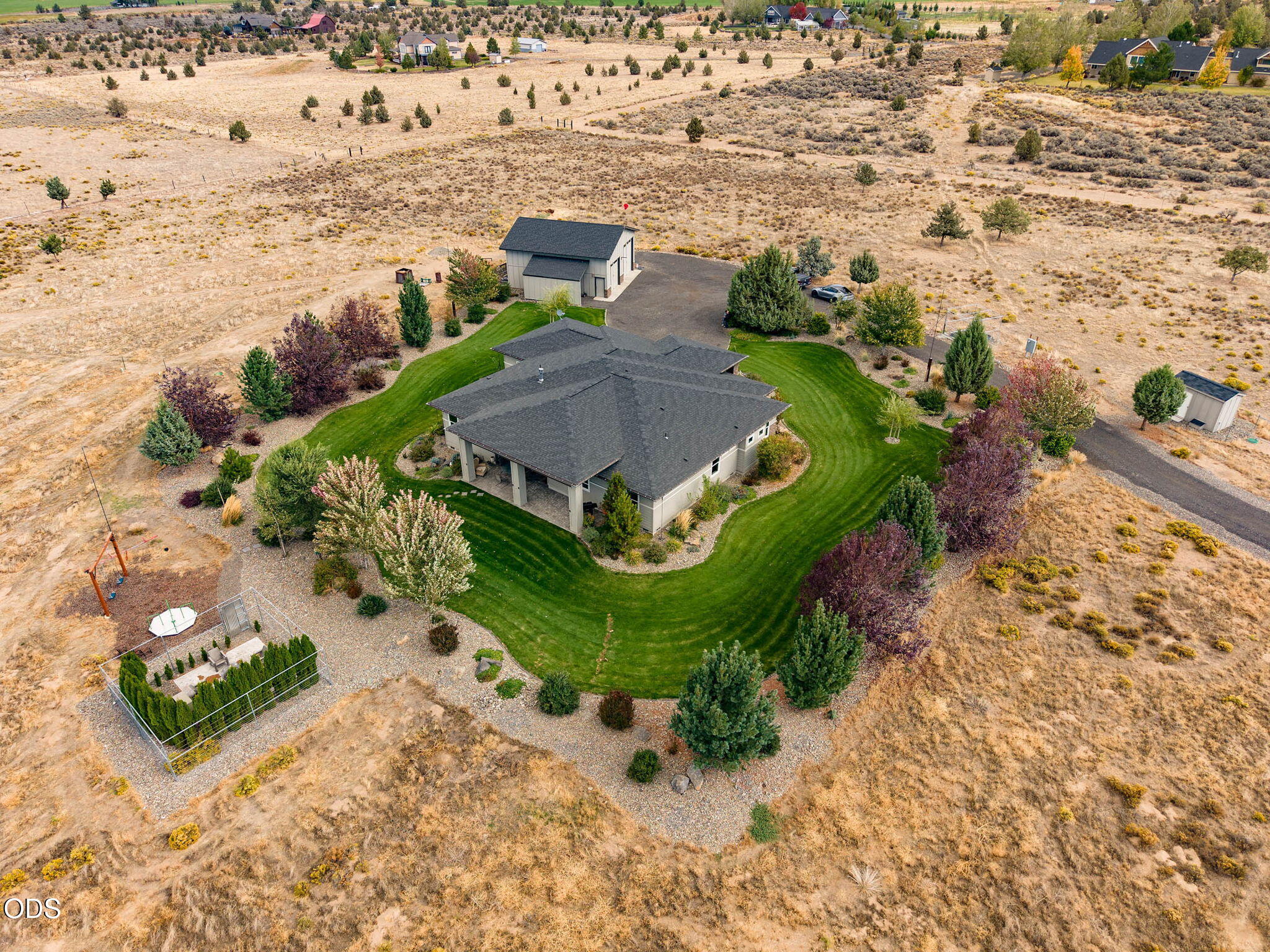 Aerial view of a home surrounded by a curved green lawn, landscaped trees, and dry open land.