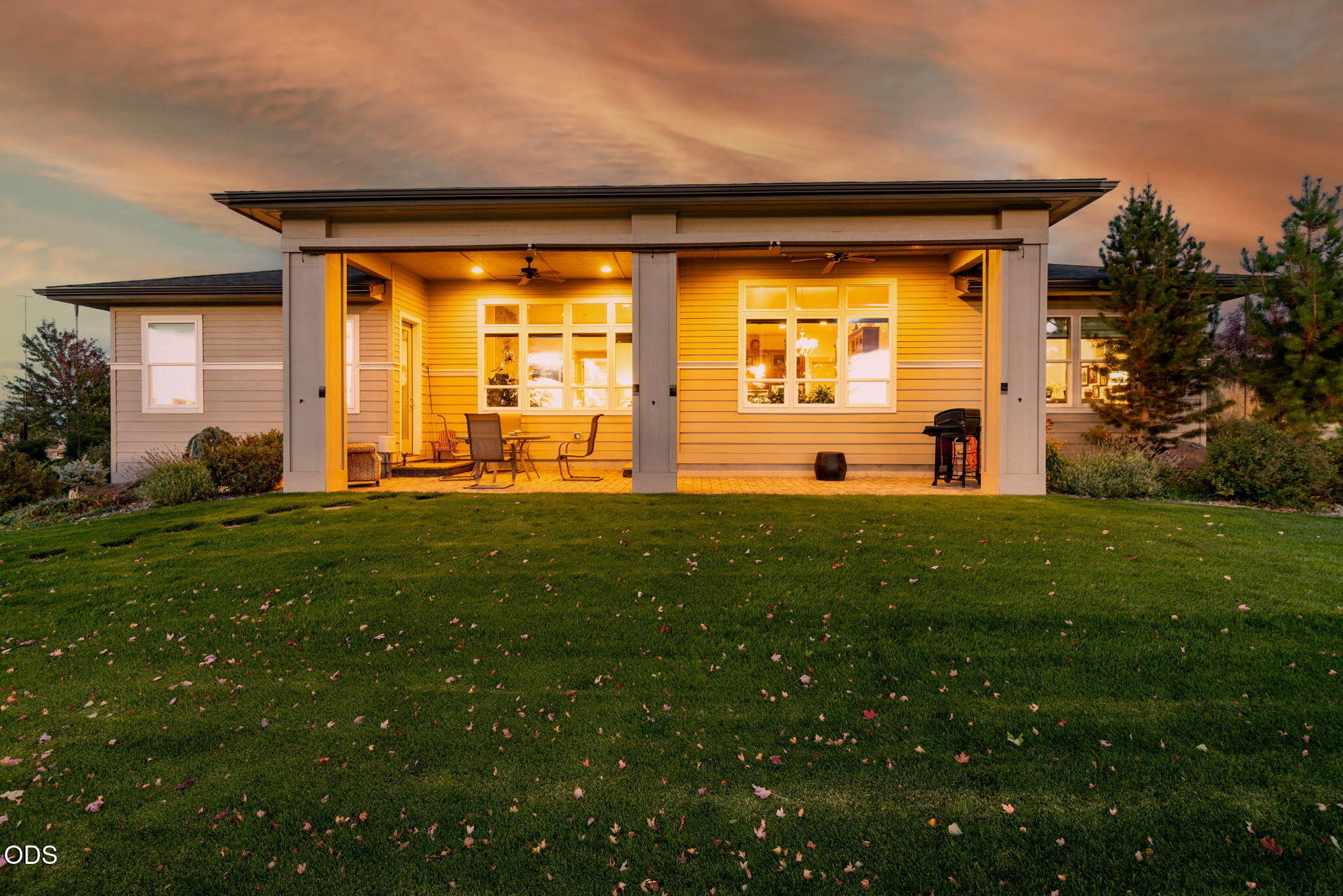 Home exterior with an illuminated covered patio, outdoor furniture, and a broad lawn at dusk.