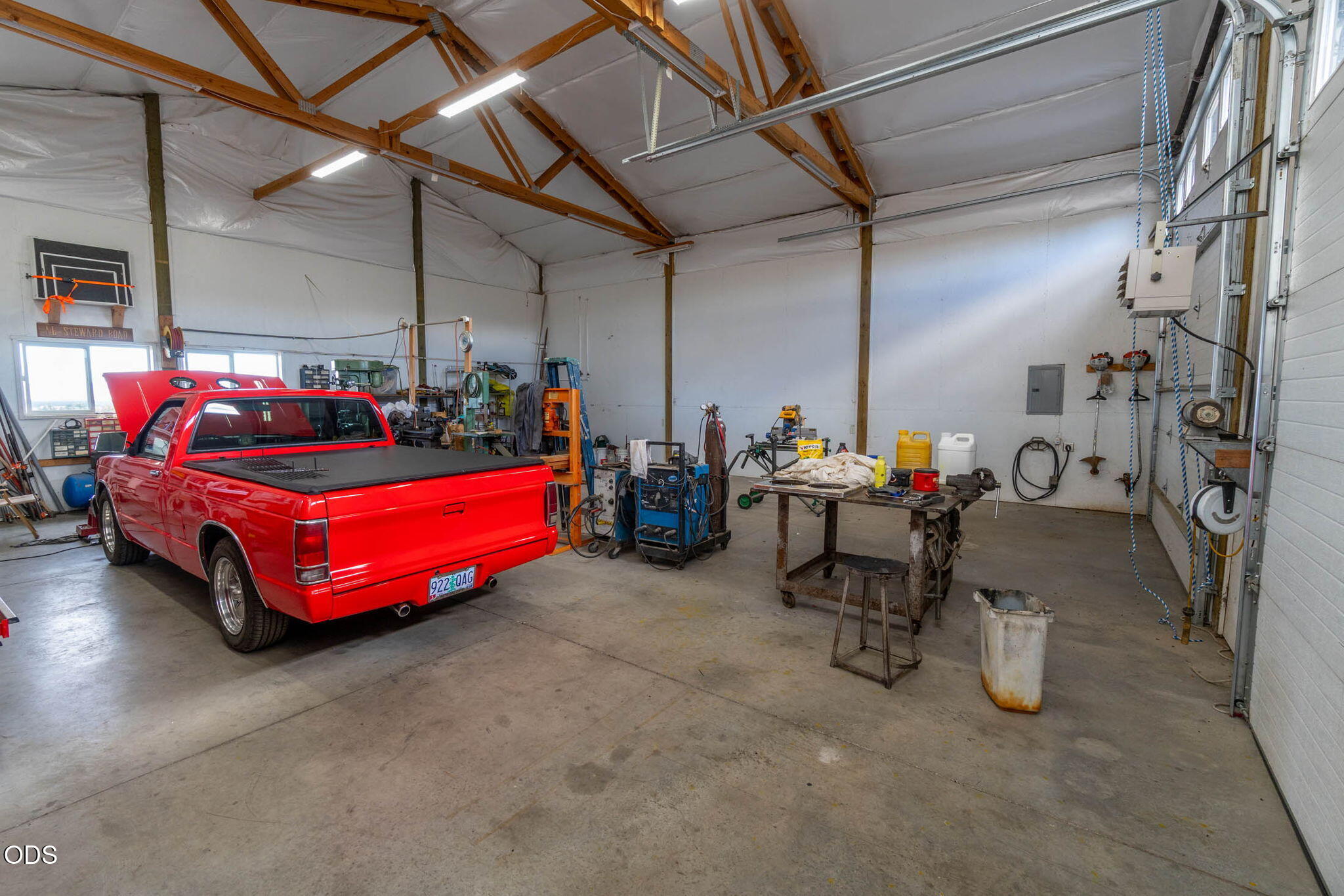 Workshop garage with a red pickup, workbenches, tools, and exposed wooden roof trusses.