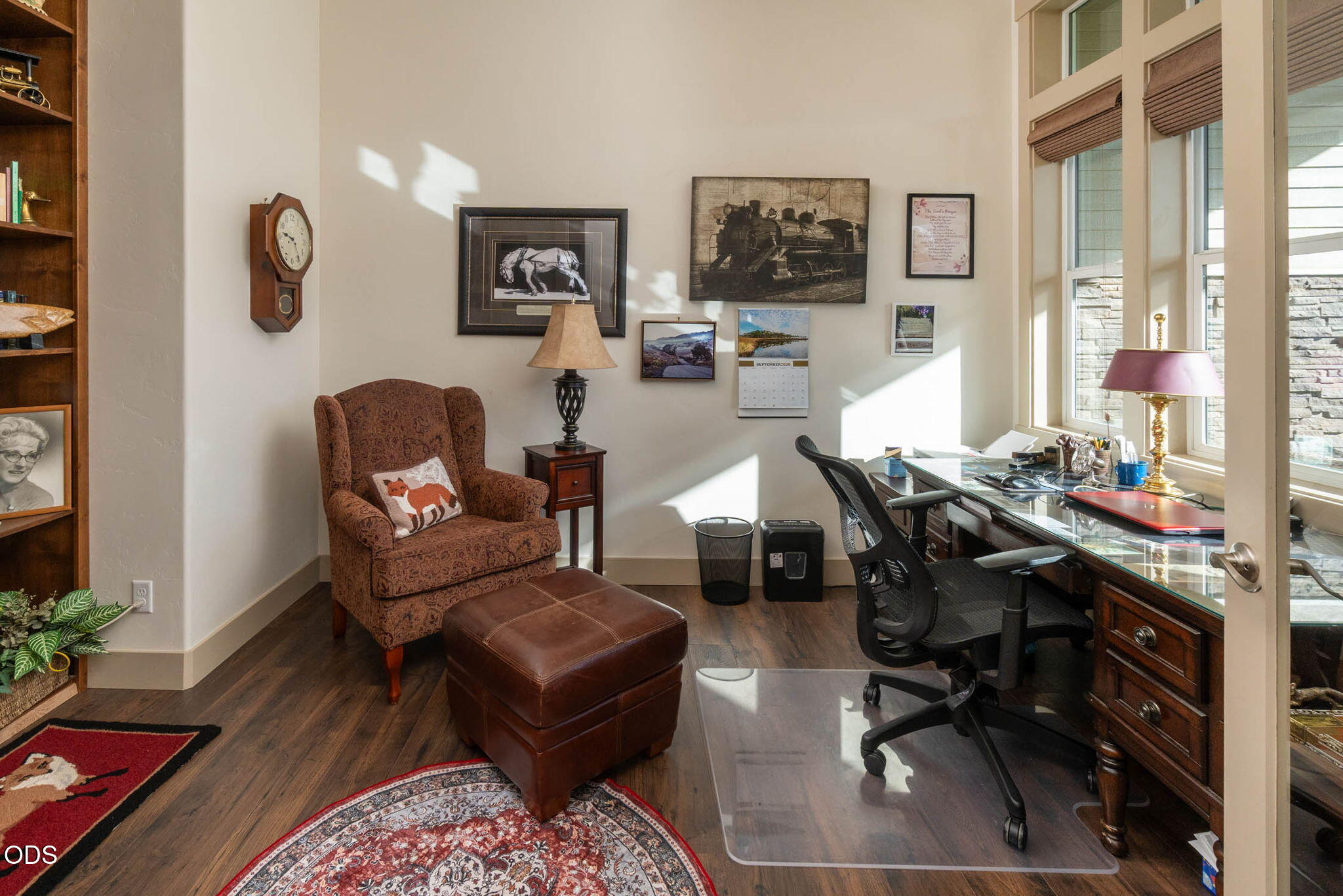 Furnished home office with a glass-topped desk, tall windows, patterned armchair, and leather ottoman.