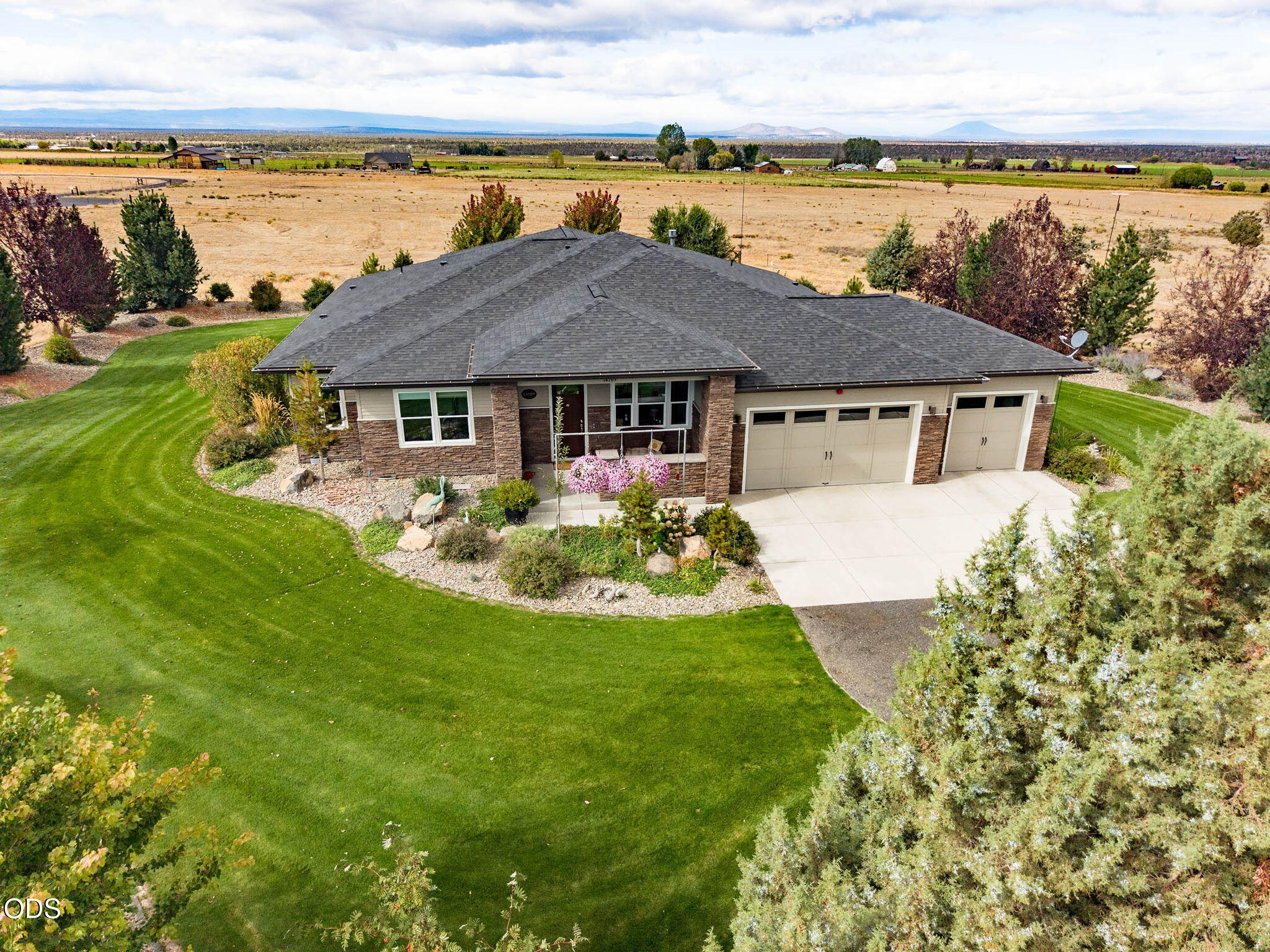 Aerial view of a stone-accented home with a three-car garage, landscaped lawn, and distant mountains.